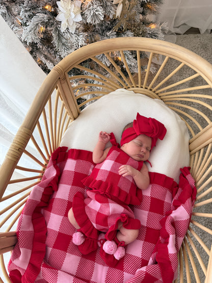 A newborn baby wearing a red and pink gingham outfit with a matching headband, sleeping in a rattan baby chair with a white cushion and a red and pink gingham blanket. A decorated Christmas tree is visible in the background.