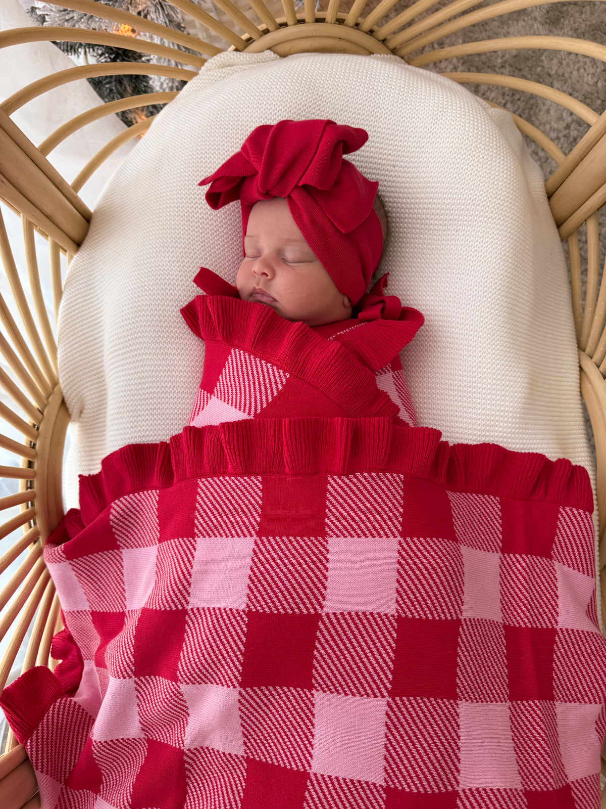 A sleeping newborn baby wearing a matching red knit swaddle blanket and red bow headband, nestled in a beige woven bassinet with a cream pillow.