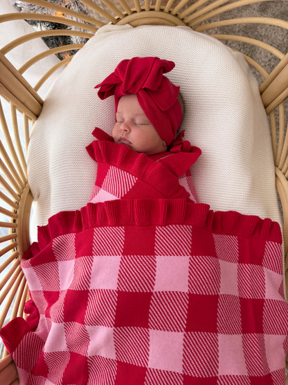 A sleeping newborn baby wearing a matching red knit swaddle blanket and red bow headband, nestled in a beige woven bassinet with a cream pillow.
