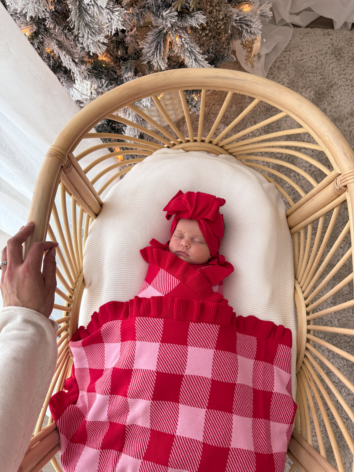 A newborn baby wearing a red bonnet and a red and pink buffalo check blanket, sleeping in a rattan bassinet with a white cushion. A hand is visible on the left side of the bassinet, and a decorated Christmas tree is in the background.