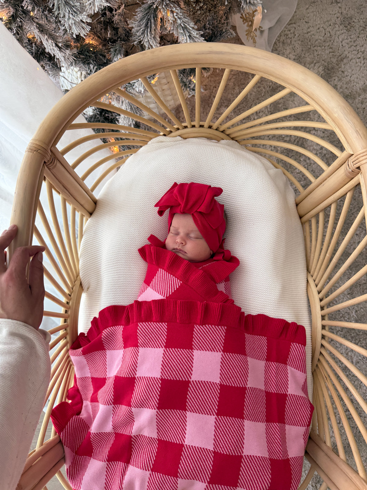 A newborn baby is swaddled in a red and pink gingham blanket and wearing a matching red bow headband, resting inside a light wood rattan bassinet with a white cushion.