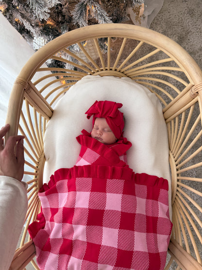 A newborn baby is swaddled in a red and pink gingham blanket and wearing a matching red bow headband, resting inside a light wood rattan bassinet with a white cushion.