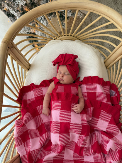 A sleeping newborn baby wearing a red ruffled bonnet and a red and pink buffalo check blanket, lying in a rattan bassinet.