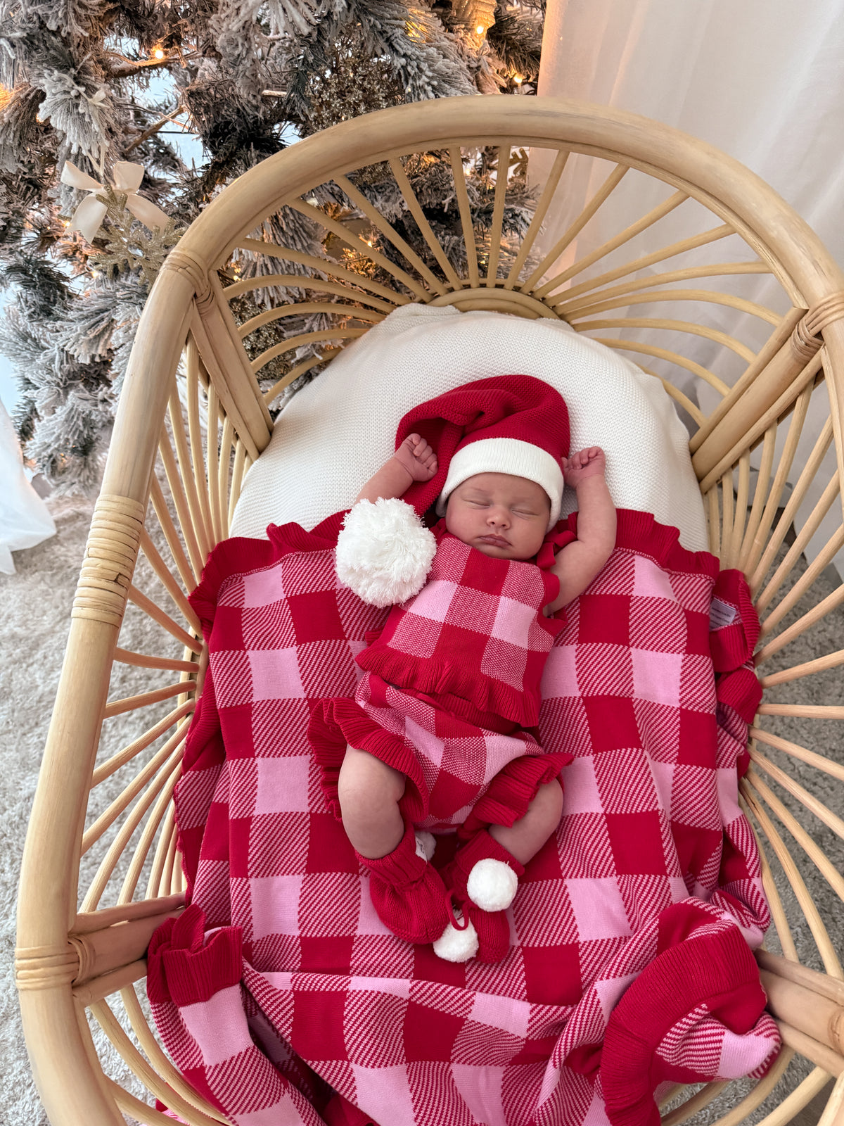 A sleeping newborn baby wearing a red and white Santa hat, matching red and white ruffled outfit, and red-and-white plaid blanket, resting inside a round rattan bassinet.