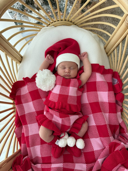 A sleeping newborn baby wearing a red and pink checkered romper, matching hat, and booties, lying on a red and pink checkered blanket inside a rattan baby chair.