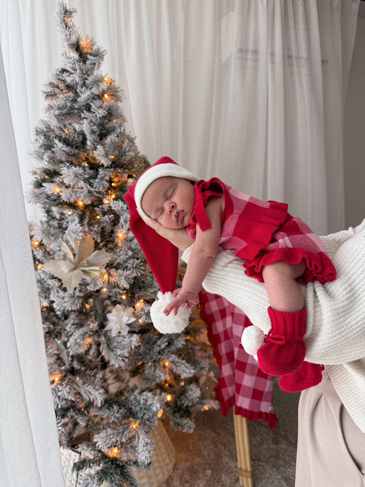 A sleeping newborn baby wearing a red and white plaid outfit, matching hat, and booties, resting on a white knit blanket next to a decorated flocked Christmas tree.