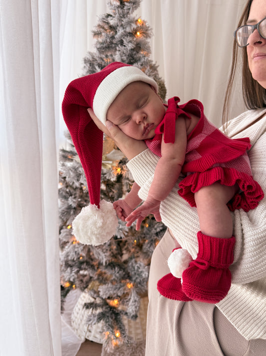 A sleeping newborn baby wearing a red and white knit Santa hat, matching red ruffled romper, and red knit booties, held by a woman in a cream knit sweater in front of a decorated Christmas tree.